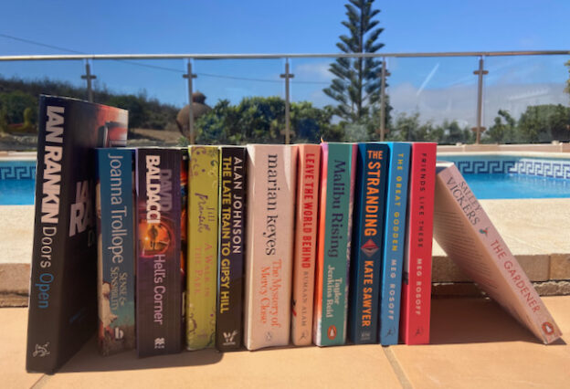 A line of books beside an outdoor swimming pool in the sun. Ian Rankin Doors Open, Joanna Trollope Sense & Senisblity, David Baldacci Hell's Corner, Jill Mansell A walk in the park, Alan Johnson The las train to gypsy hill. Marian Keye The mystery of mercy close, Rumaan Alam's Leave the world behind, Malibu Rising by Taylor Jenkins Reid, The Stranding by Kate Swayer, The Great Godden and Friends like these both by Meg Rosoff and Salley Vickers The Gardener