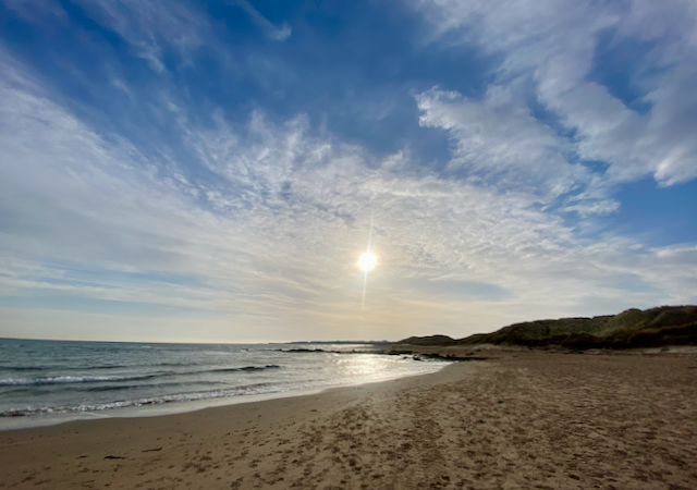 Image of the beach at Seahouses in Northumberland on a bright blue sky sunny February day. The sun is partially covered by a streak of white cloud, the sea to the left of the image, sand in front and to the right,