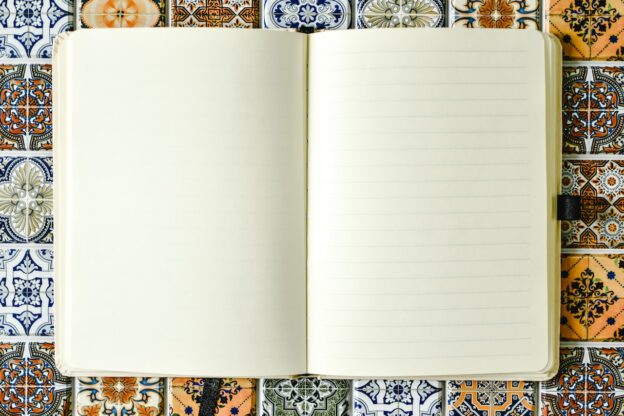 Image is of an open notebook with two facing blank pages. It's sitting on a tile table top, with different mediterranean patterned tiles