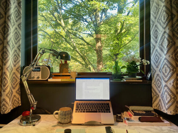Harriet's desk covered in notebooks, her laptop, pens and pencils and an anglepoise lamp. Behind is a windoow full so sky and green leaf trees and a window sill with ceramic jugs, a radio, a white flowered plan and a vase full of pencils.