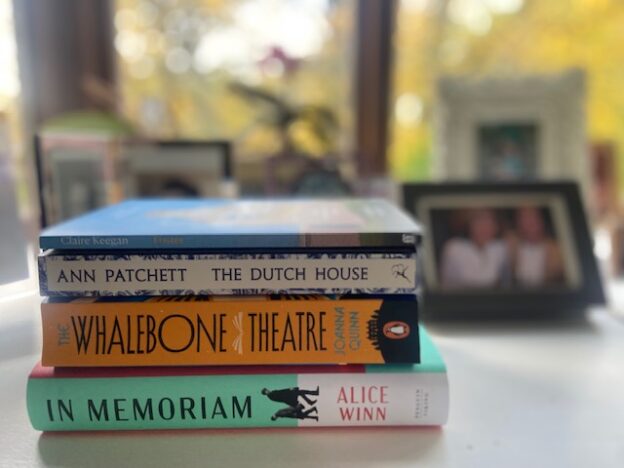 A pile of books on a window sill surrounded by photo frames. The books are: The Whalebone Theatre by Joanna Quinn. In Memorium by Alice Winn. The Dutch House by Ann Patchett. Tomorrow and tomorrow and tomorrow by Gabrielle Zevin. Foster by Claire Keegan
