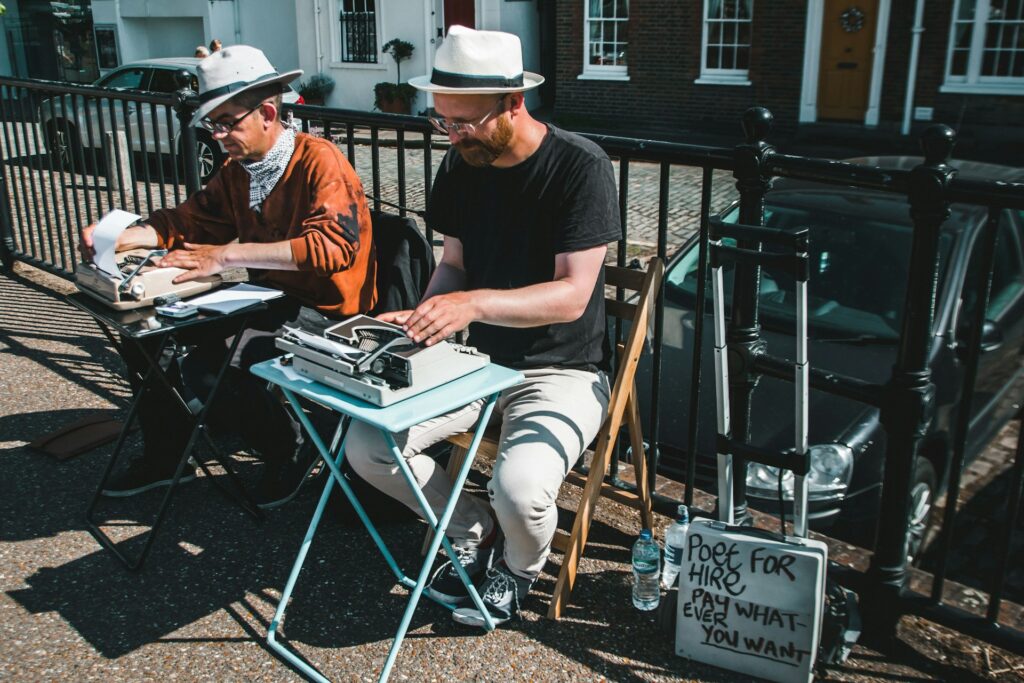 Two men sat on the pavement wearing white trilby hats with a black ribbon, sitting on wooden folding chairs with typewriters on little metal folding tables in front of them. Windows and buildings and railings behind them