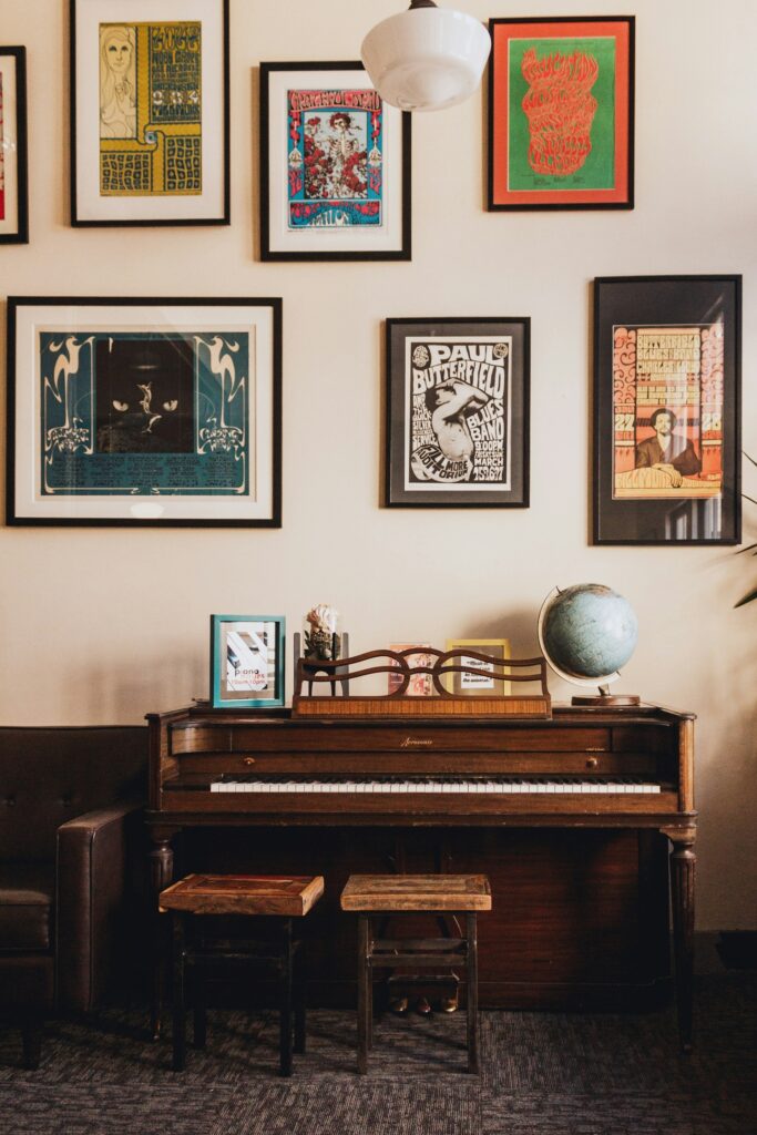 A dark brown wood piano is set against a cream, pale painted wall with paintings and pictures on it. The piano has a globe on the right handside and two piano stools. It's a traditional looking piano.