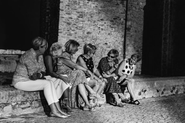 Black and white photo of a group of six women sitting on a low stone wall heads turned to each other in what looks like amiable conversation