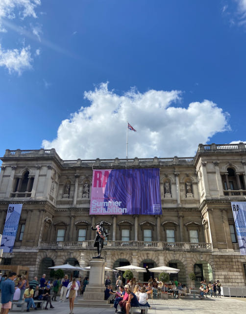 Image of the exterior of the Royal Academy of Art in London against a blue sky and white clouds. It's a grand building, old with two stories, pale stonework with pillars.